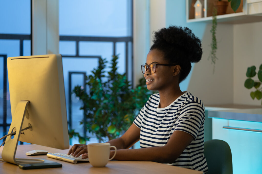 Young happy african woman freelancer in glasses working remotely in cozy home office with houseplant on background, remote female developer coding on computer, enjoying flexible freelance schedule